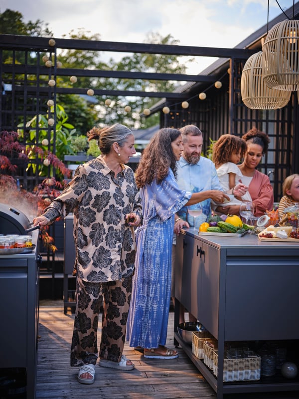 A group of people of all ages in an outdoor kitchen party setting around a BÅTSKÄR charcoal bbq and closed kitchen unit.