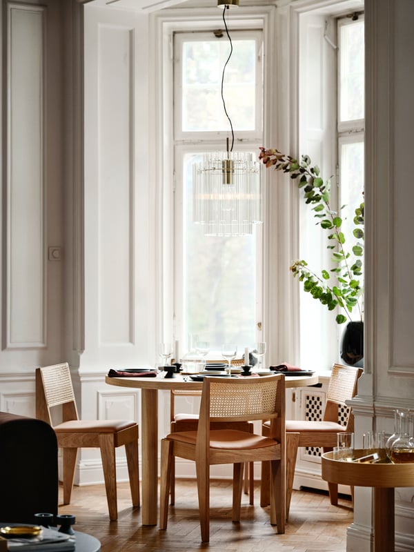 Scena di sala da pranzo con lampada a sospensione STOCKHOLM 2025 placcata in ottone con tubi di vetro sospesi sopra un set da pranzo con tavolo in rovere e sedie in rovere e rattan.