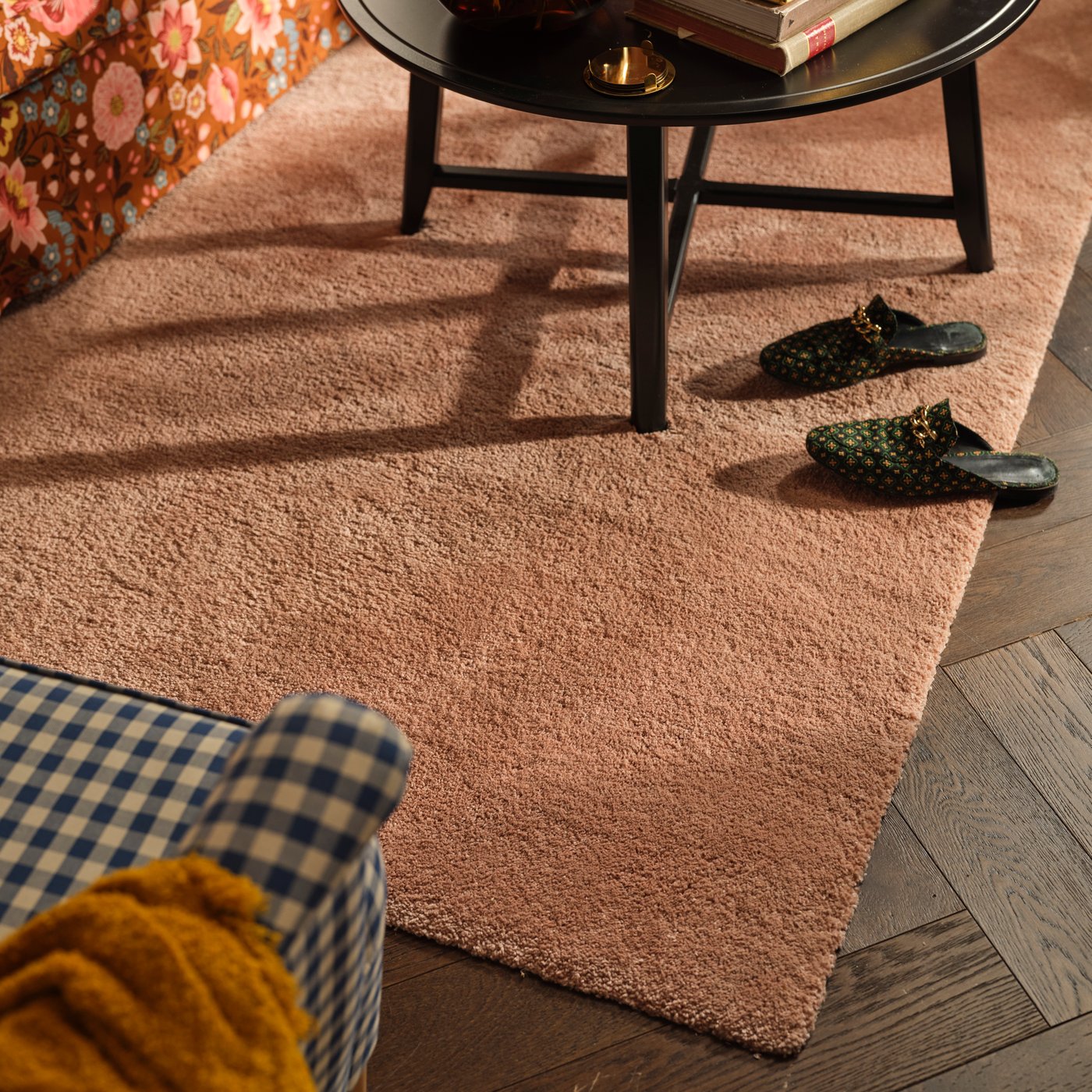 A STOENSE rug in pale pink is on a living room floor. It has a black coffee table on top and a chequered armchair next to it.