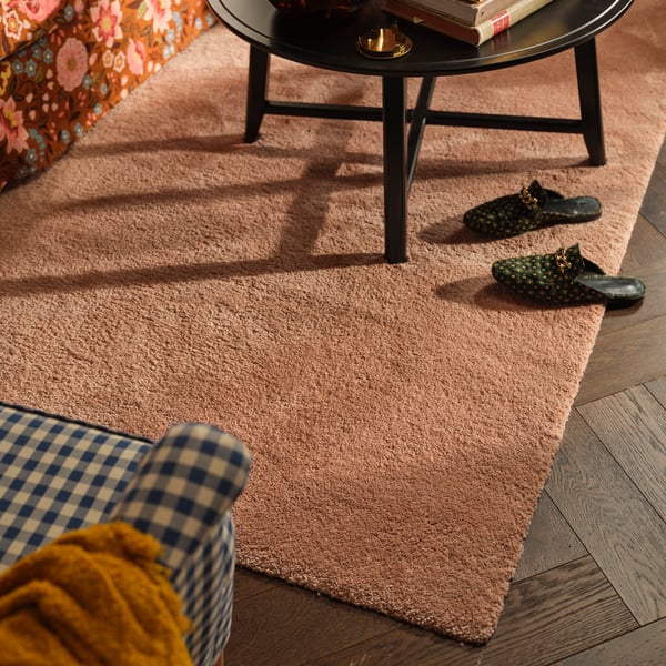 A STOENSE rug in pale pink is on a living room floor. It has a black coffee table on top and a chequered armchair next to it.