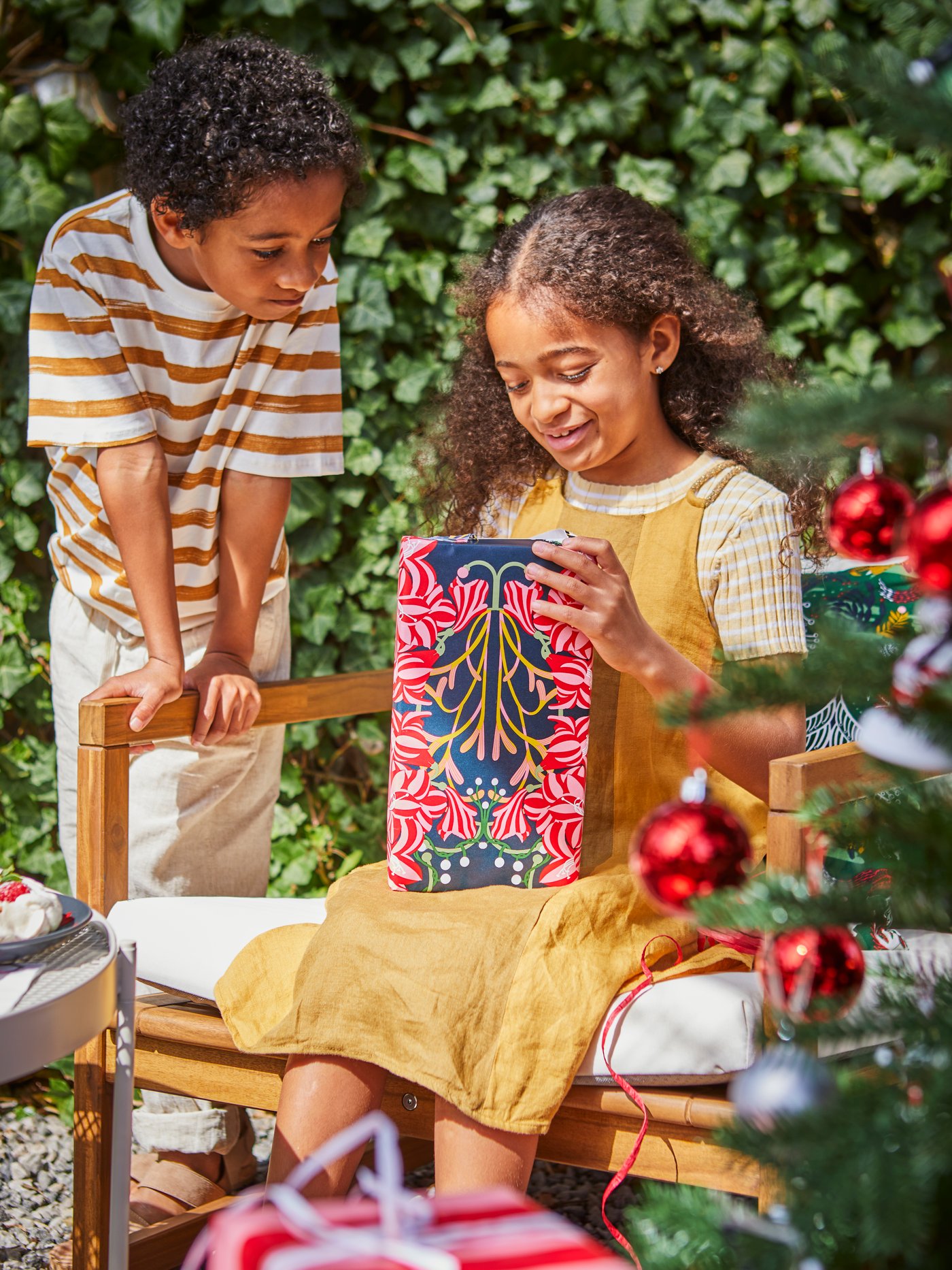 Unos niños abriendo dos paquetes navideños al lado de un árbol de Navidad.