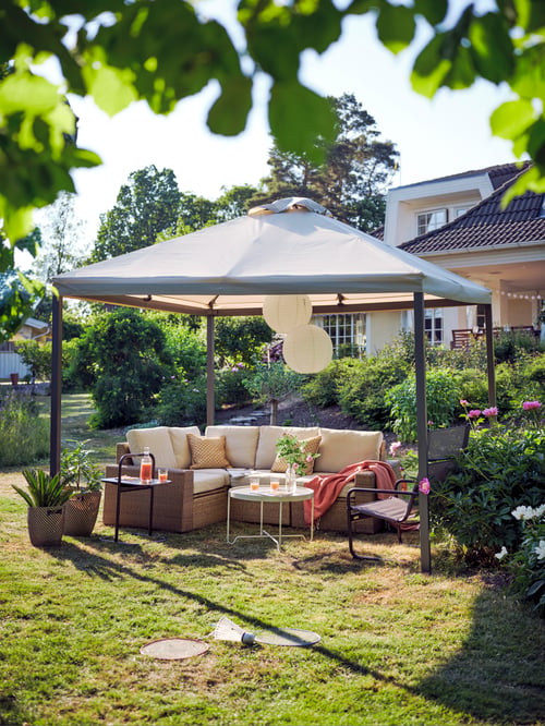 Ein SOLLERÖN Ecksofa für außen mit beigen Kissen und ein schwarzer Sessel unter einem HIMMELSÖ Pavillon auf einer sonnigen Gartenwiese.