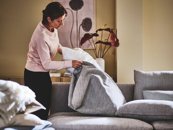Person arrangerar kuddar på grå soffa i vardagsrum med tavla och växter som dekoration.