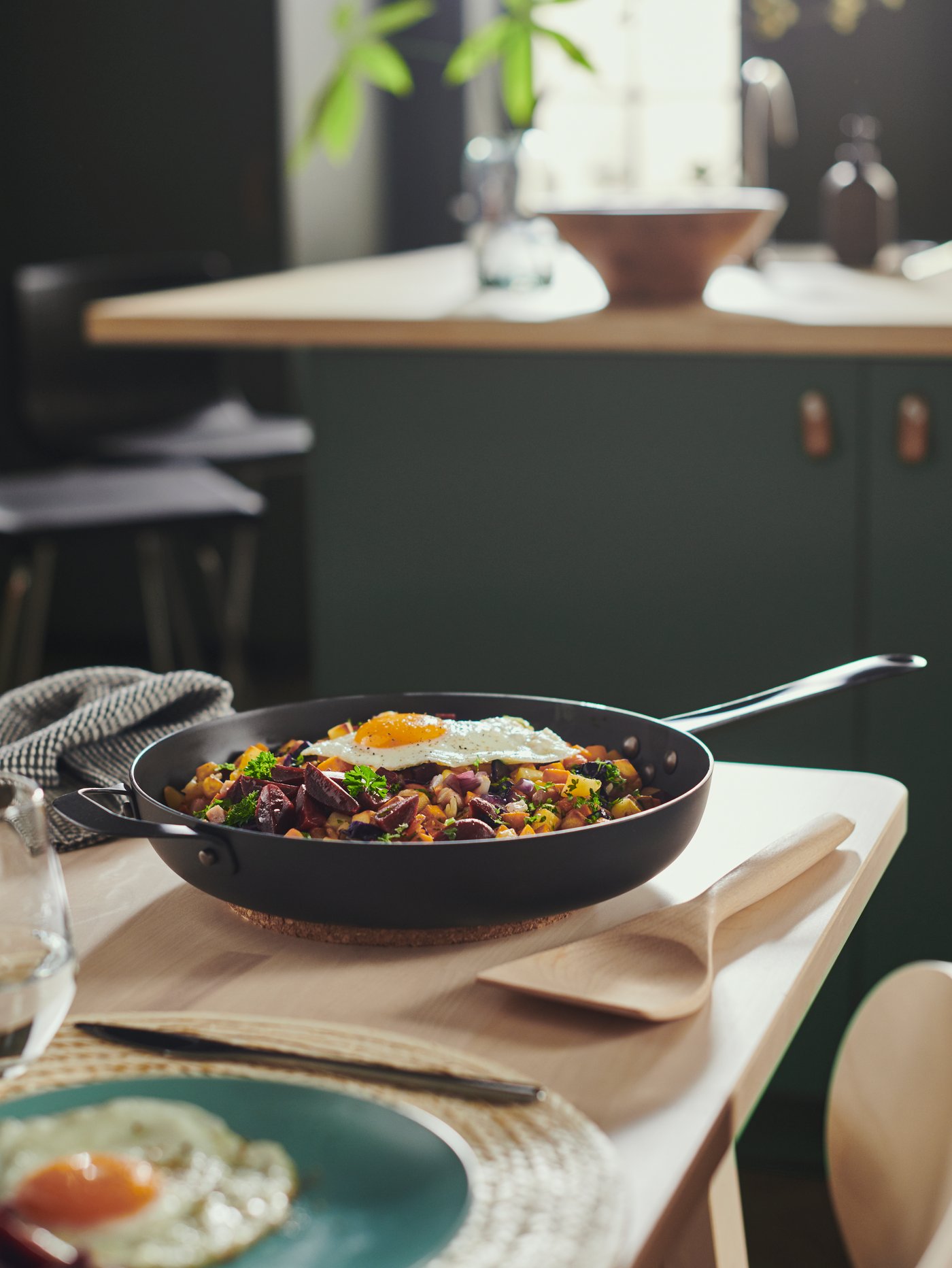 A VARDAGEN frying pan with pyttipanna and a beech wok spatula on a birch table set with light turquoise FÄRGKLAR plates.