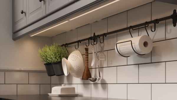 IKEA MITTLED LED bänkbelysning som styrs trådlöst sitter under överskåpen i ett kök. Under sitter en väggförvaring där det hänger olika köksredskap.