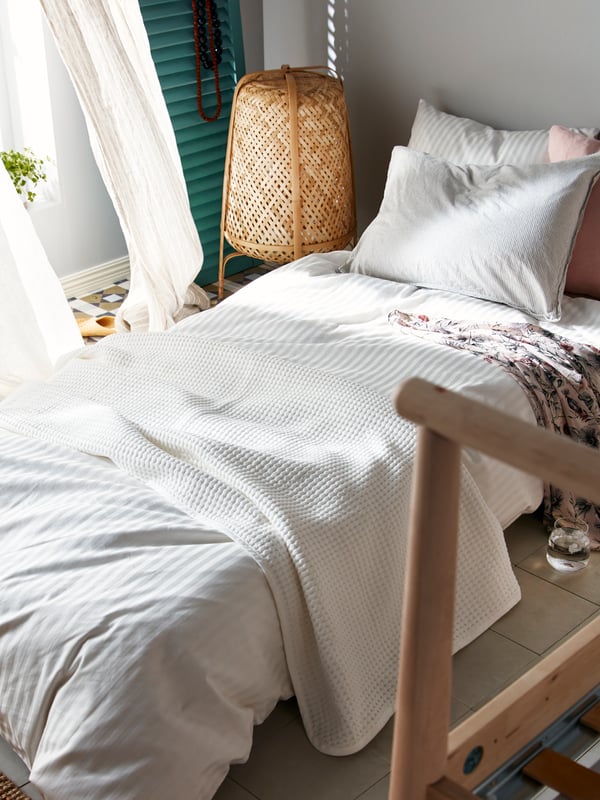 A bed with VÅRELD bedspread and NATTJASMIN quilt cover and pillowcase in white, with a KNIXHULT floor lamp and curtains beside.