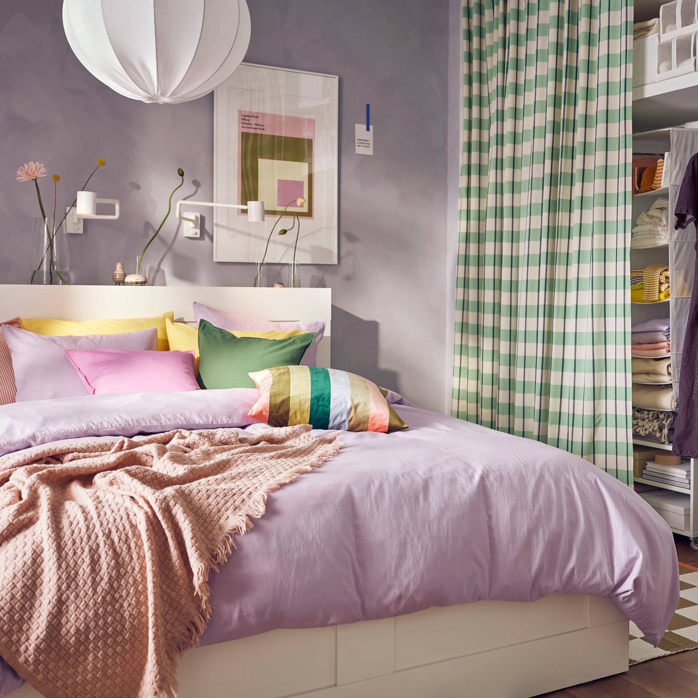 In una stanza lilla, un letto BRIMNES con biancheria NATTSVÄRMARE lilla. Una tenda GLANSHAGTORN verde nasconde una soluzione per organizzare i vestiti.
