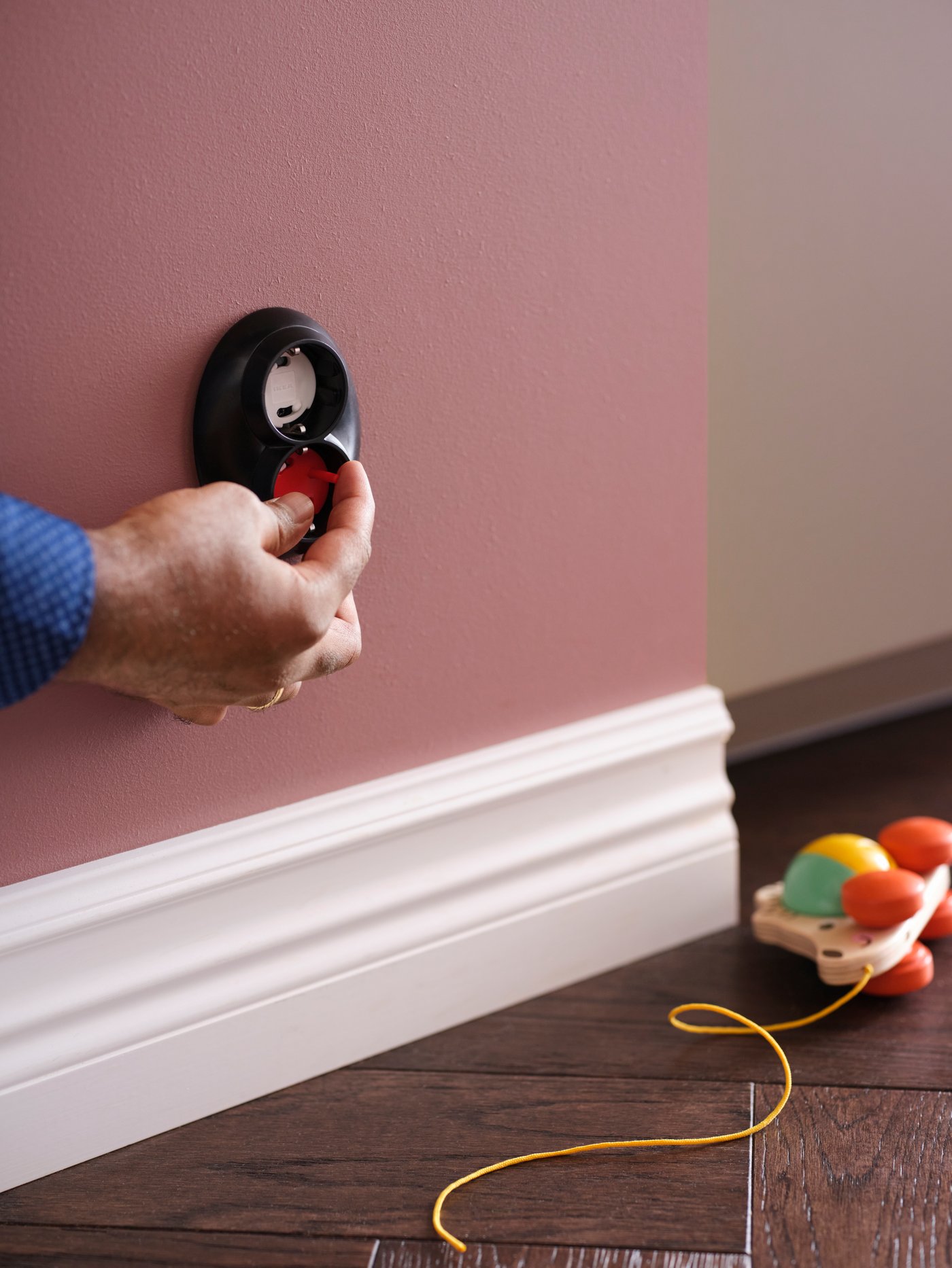 A person removing a white PATRULL safety plug from a wall socket using a coloured tool, plus a pull toy on the floor.