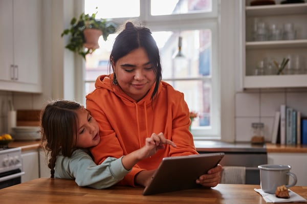 A person wearing an orange hoodie is using a tablet with a child in a kitchen