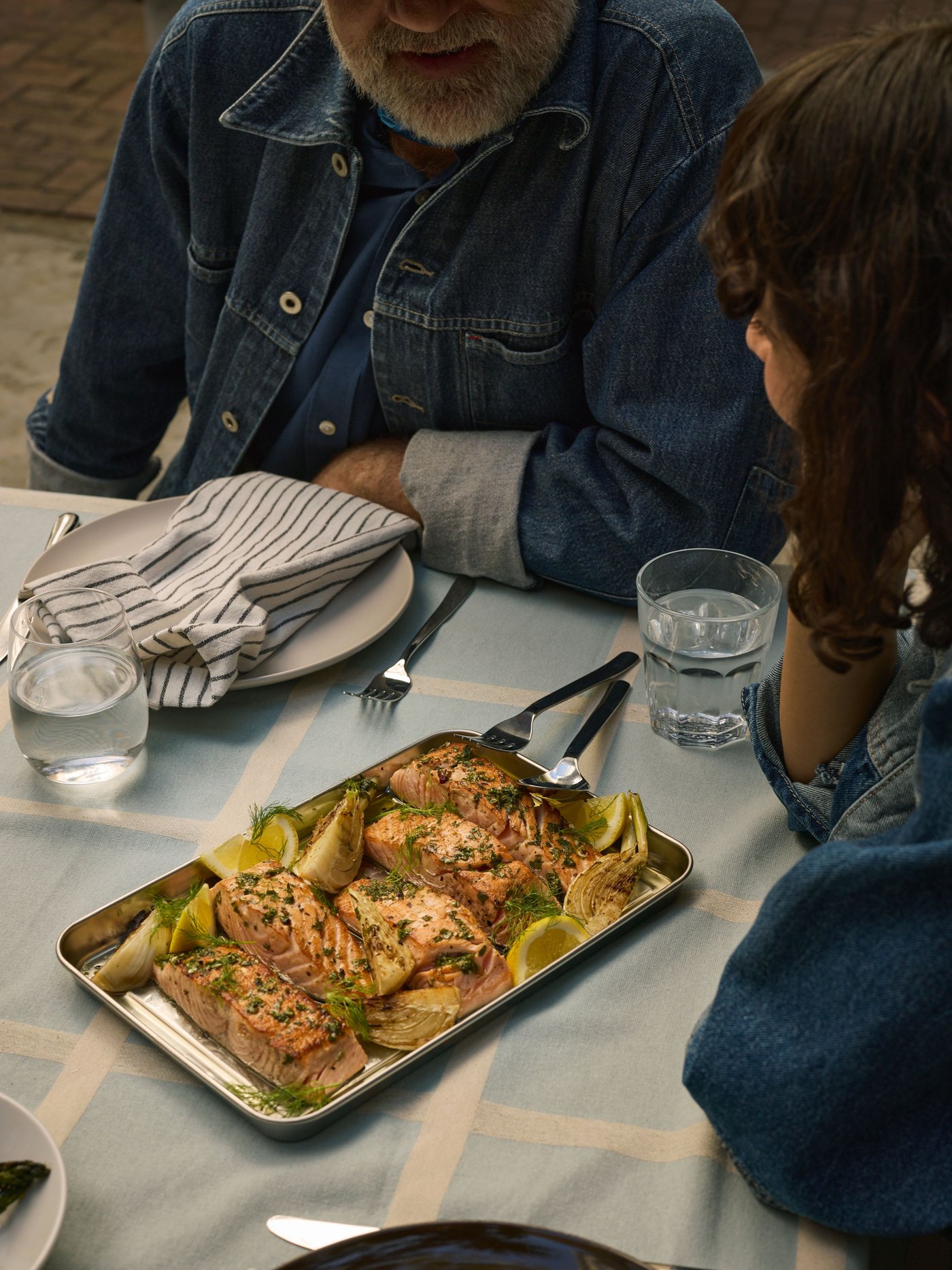 شخصان يرتديان سترات من الجينز يجلسان أمام طاولة عشاء مجهزة بانتظار مأكولات مشوية على صينية تقديم GRILLTIDER 