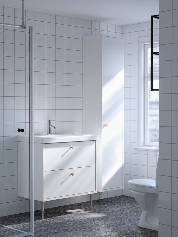A white HAVBÄCK wash-stand with two drawers and a high cabinet with a door next to it are placed in a white-tiled bathroom.
