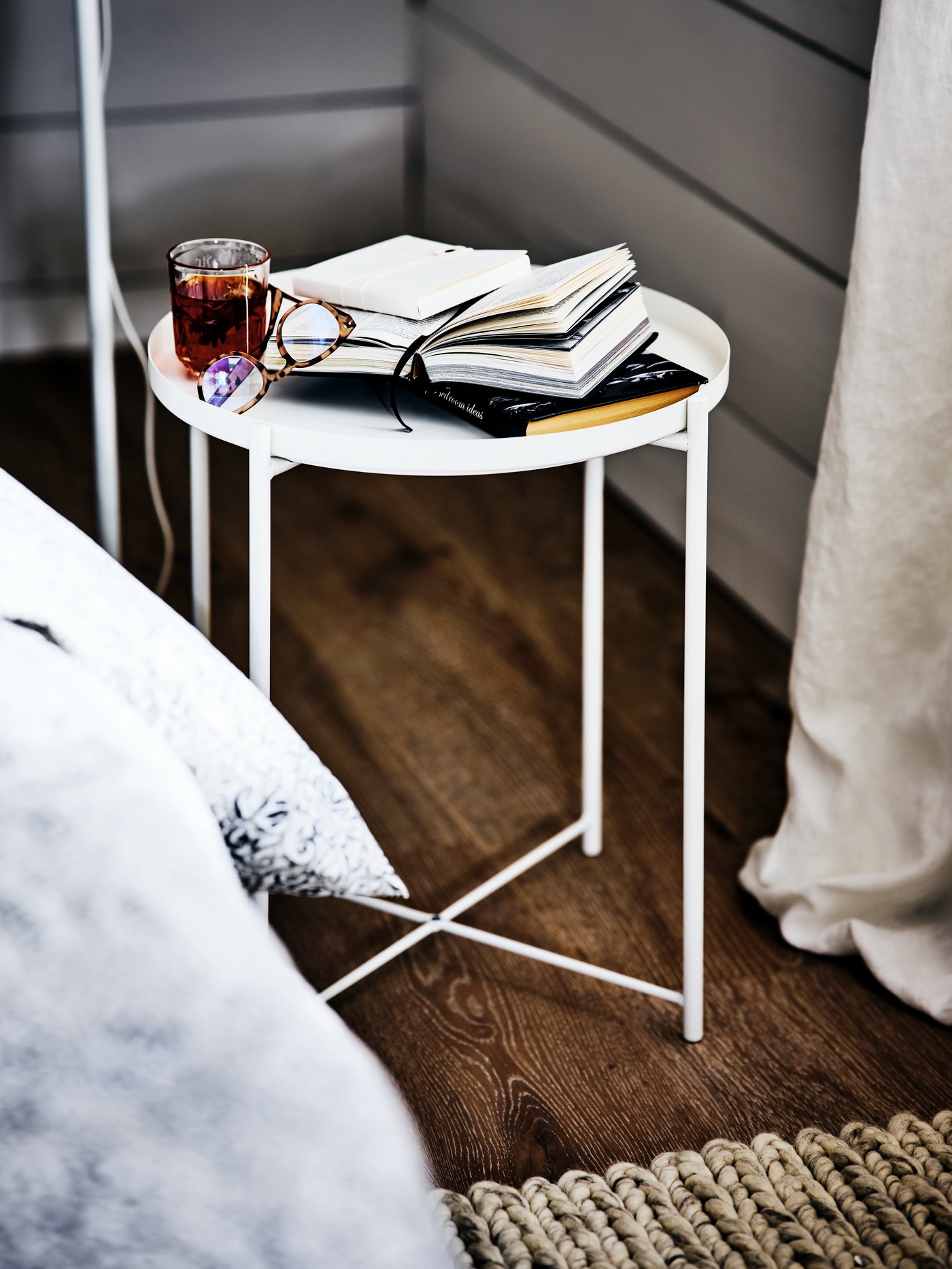 A white GLADOM coffee table in the corner of a room beside a bed, with some books, a pair of glasses and a drink on it.