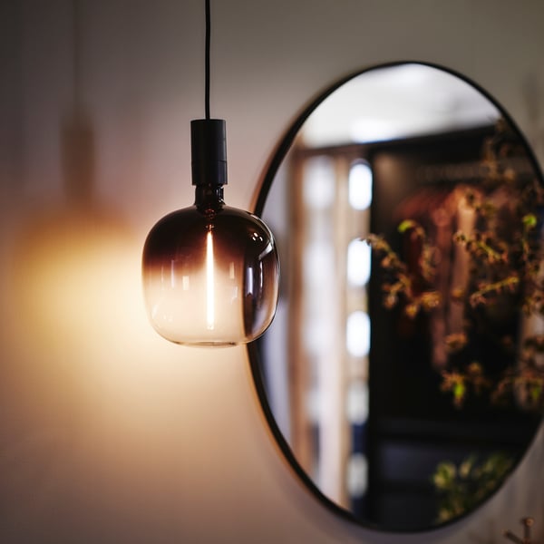 Close-up of a MOLNART light bulb with a soft light, hanging down in front of a round mirror in the background.