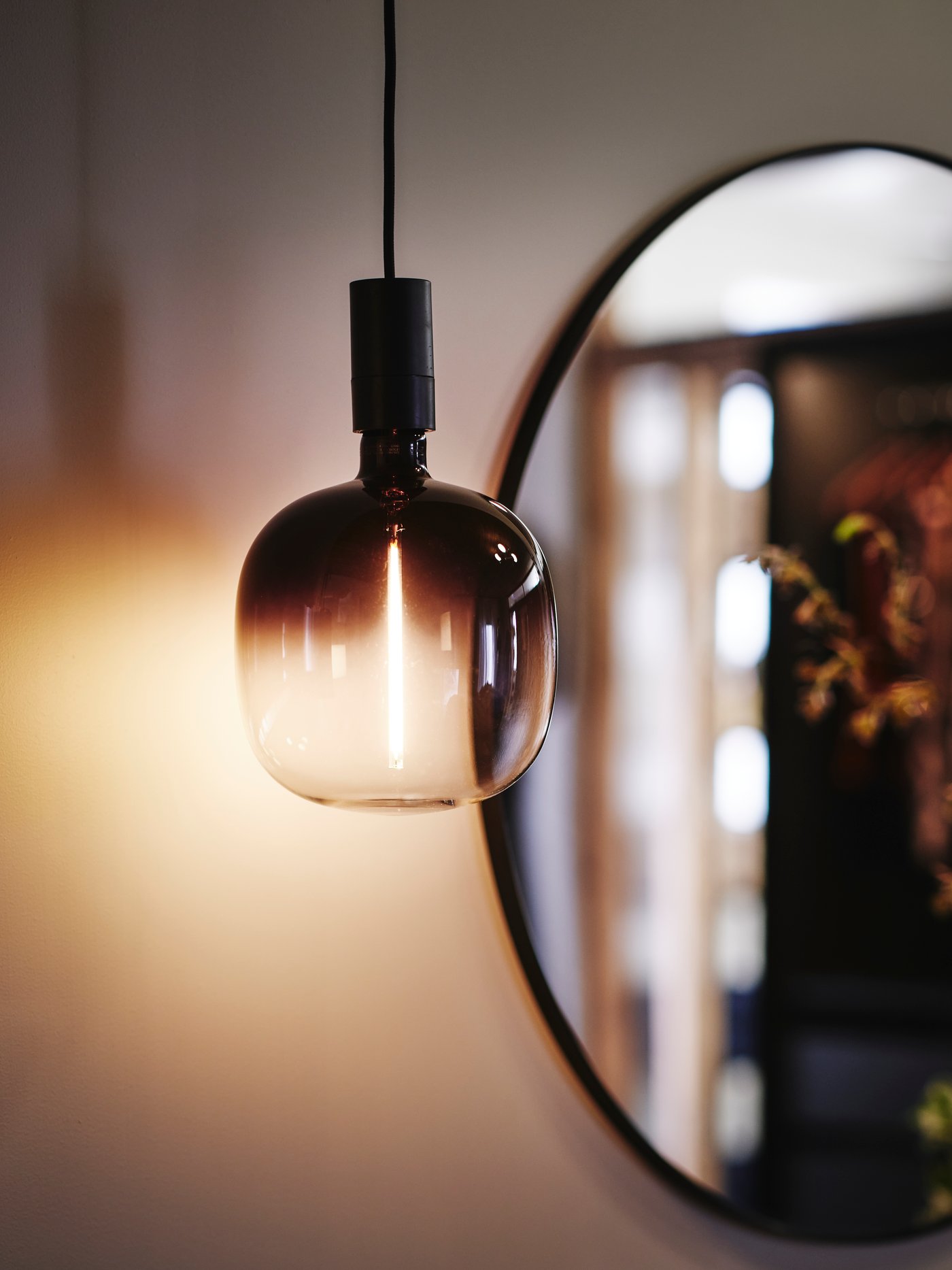 Close-up of a MOLNART light bulb with a soft light, hanging down in front of a round mirror in the background.