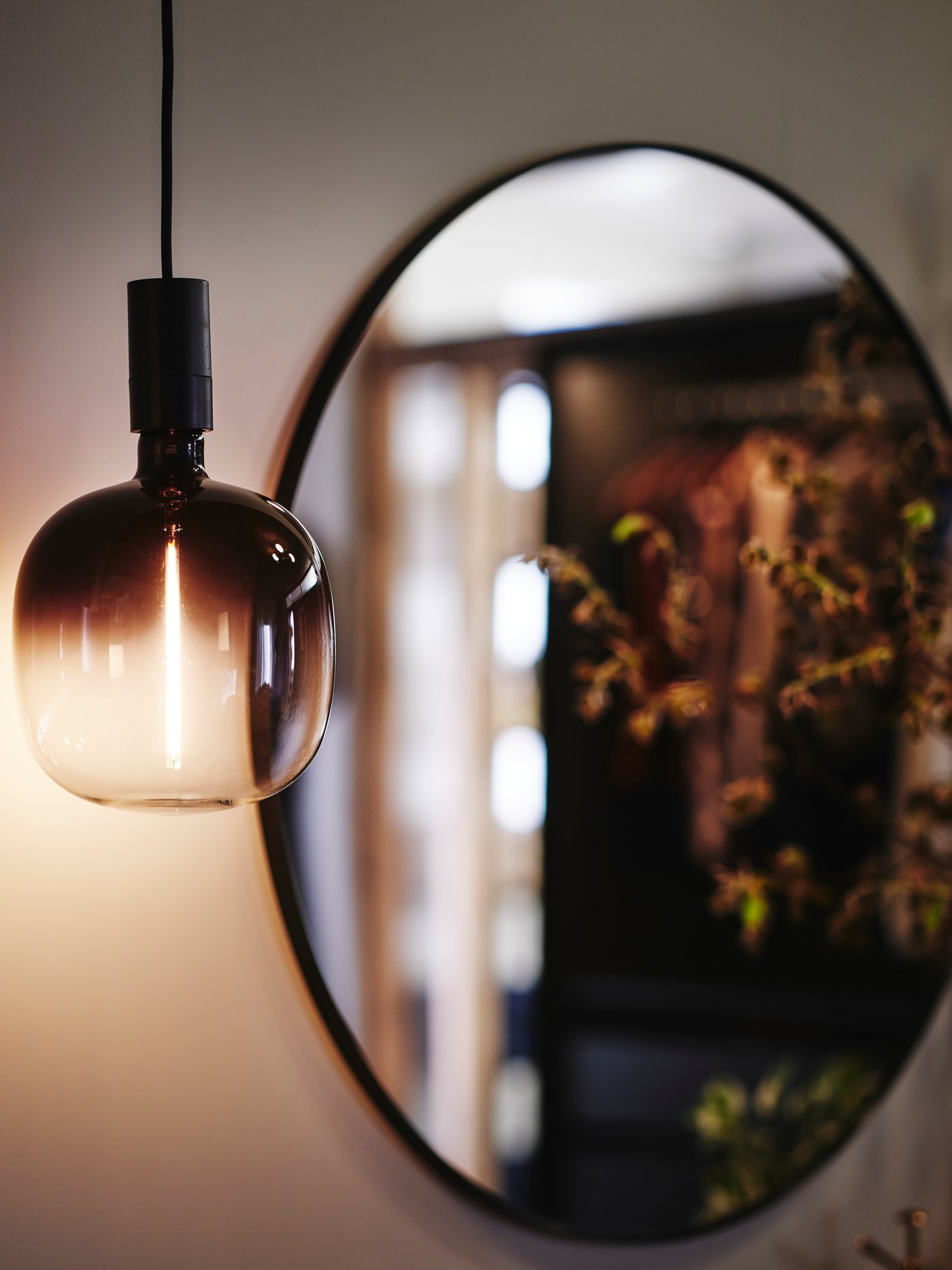 Close-up of a MOLNART light bulb with a soft light, hanging down in front of a round LINDBYN mirror in the background.