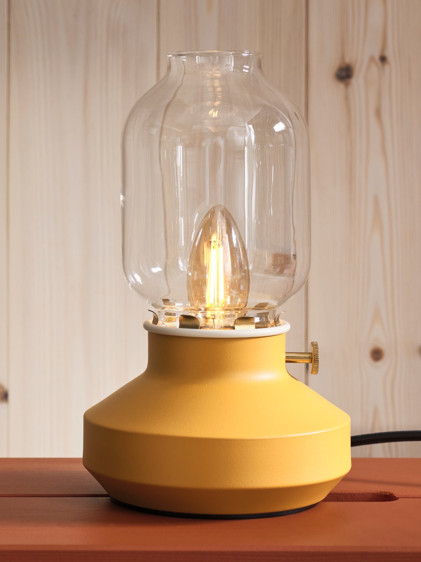Żółta lamapa TÄRNABY stojąca na czerwonym stołku na tle drewnianej ściany.