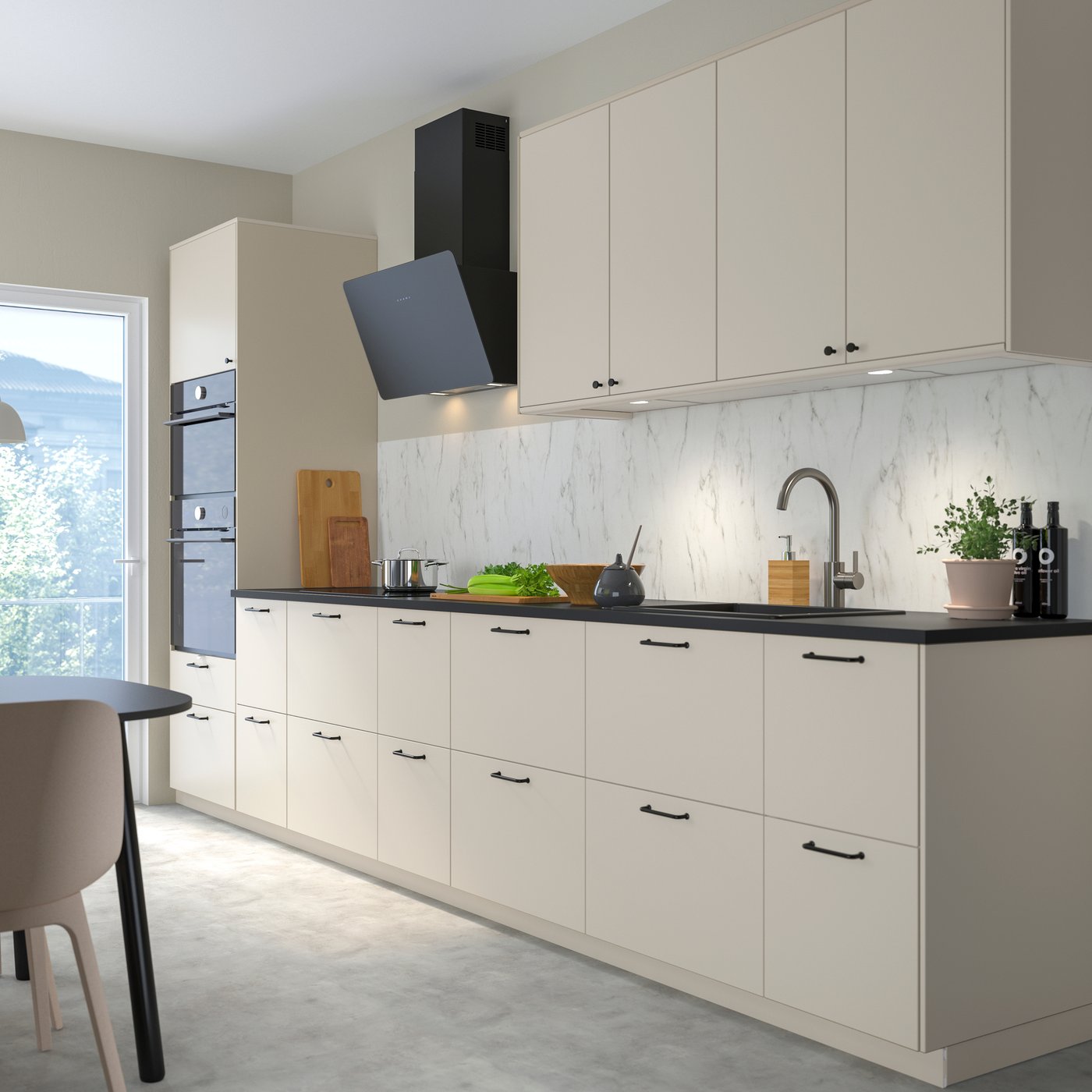 Modernt kök med beige skåp, svart bänkskiva, vit marmorbakgrund, inbyggd ugn och grönsaker på bänken.

