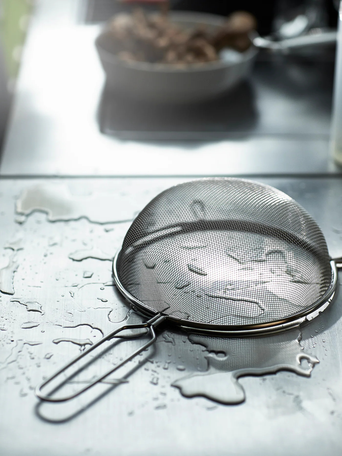 An IDEALISK strainer is placed on top of a wet stainless steel surface. In the background, there’s grilled food.
