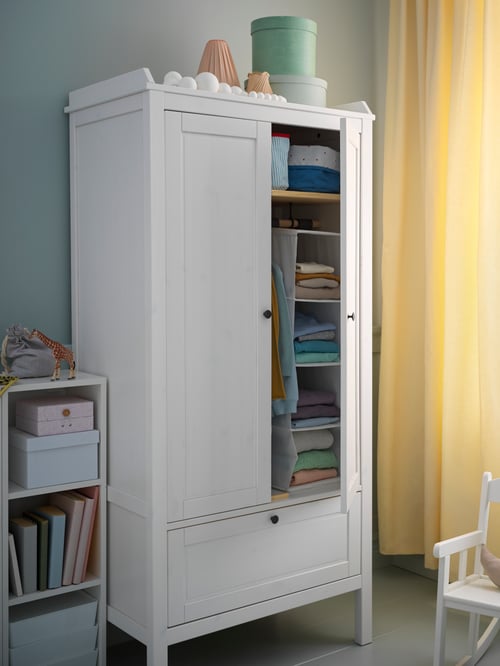 Kinderzimmer mit weißem Kleiderschrank aus Holz und einer geöffneten Tür, u. a. mit einem weißen Regal mit Kartons und gelben Gardinen.