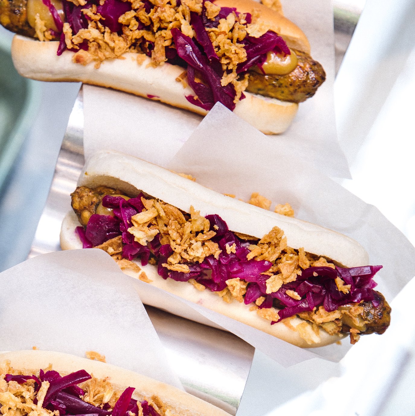 Three veggie hot dogs with pickled red cabbage, spicy mustard and fried onions waiting to be collected on a serving stand.
