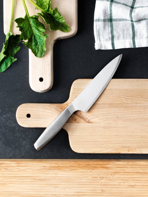 Two kitchen knives and three different cutting boards beside some vegetables and a kitchen towel on a black countertop.