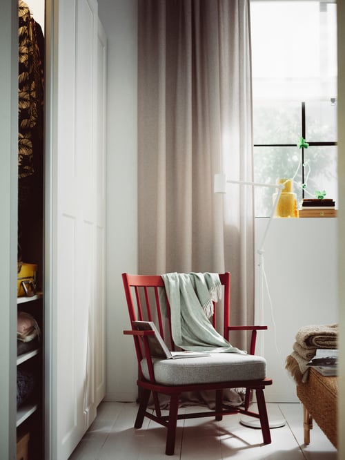 Schlafzimmer mit einem PERSBOL Sessel und einem HOLMVI Plaid in einer Zimmerecke neben einem Fenster mit Gardinen und einer NYMÅNE Stand-/Leseleuchte.