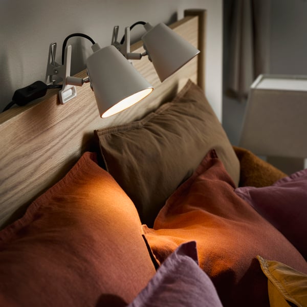 A close up of HAVSSALLAD clamp stptlight on a light wooden bed frame with pink and brown pillows below.