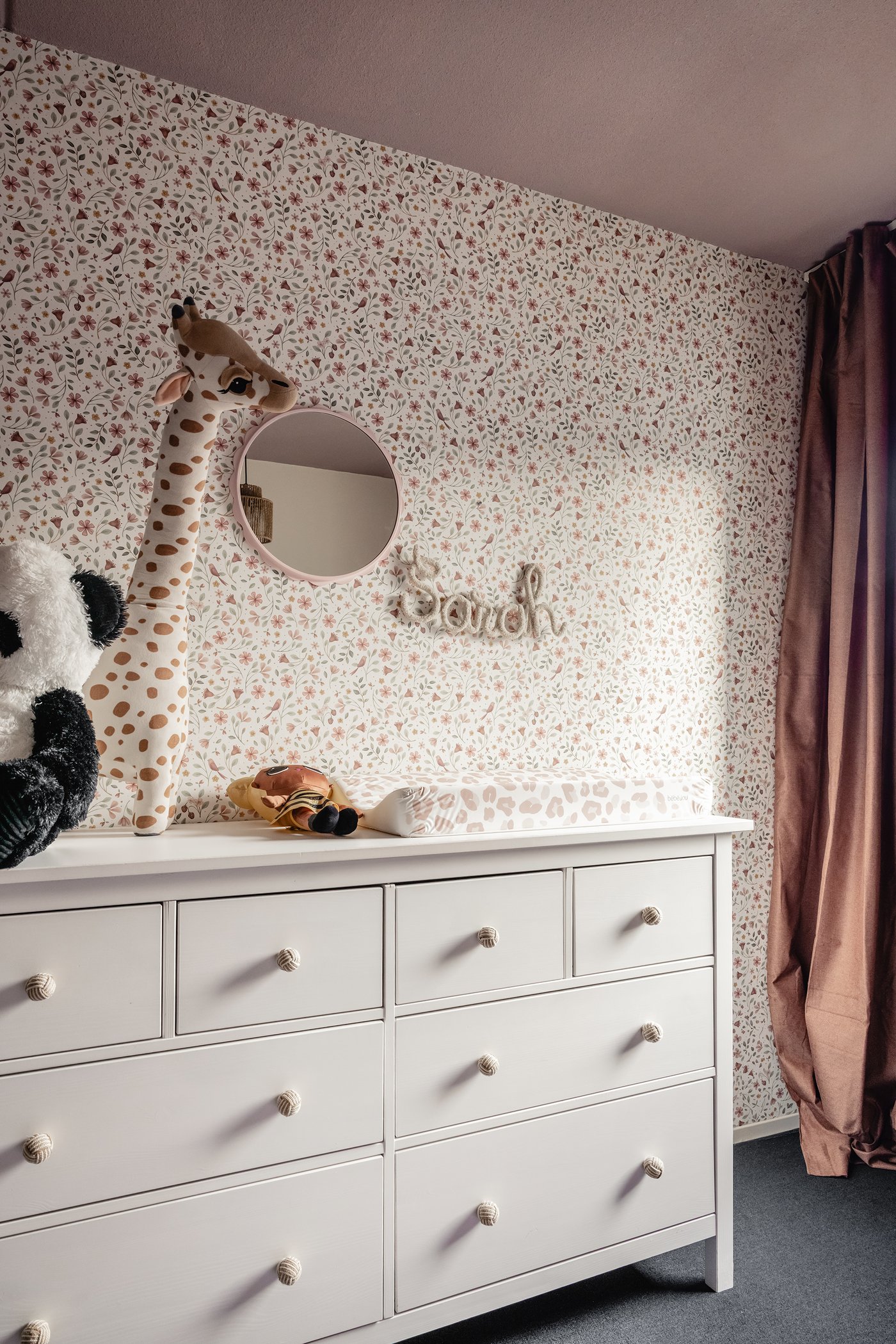 Een kinderkamer met een witte HEMNES ladekast met 8 lades. Op de kast staan een paar knuffels, aan de muur hangt een roze, ronde DAGGKAPRIFOL spiegel. Op de muur zit een behang met bloemetjes print.