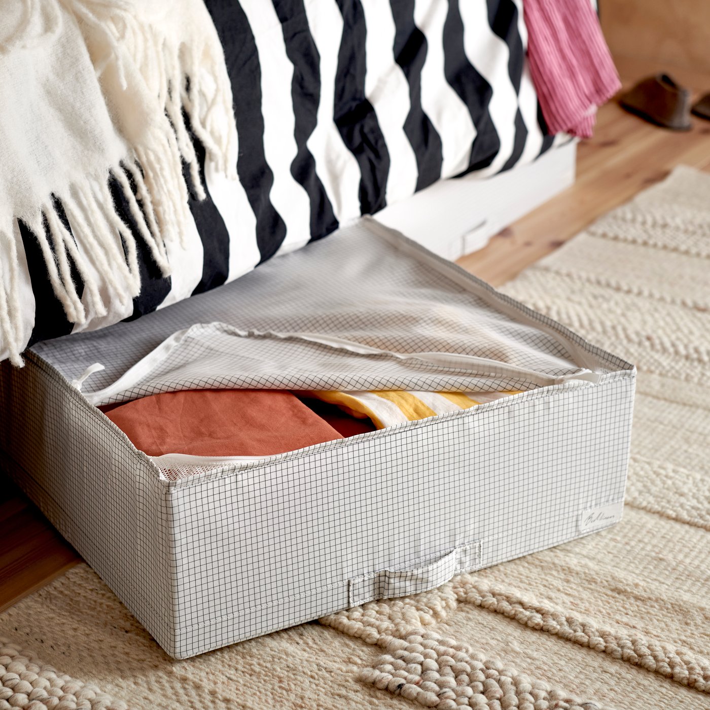 A STUK storage case with clothes in it lies on a mat on the floor, partly under a bed with a striped duvet cover.