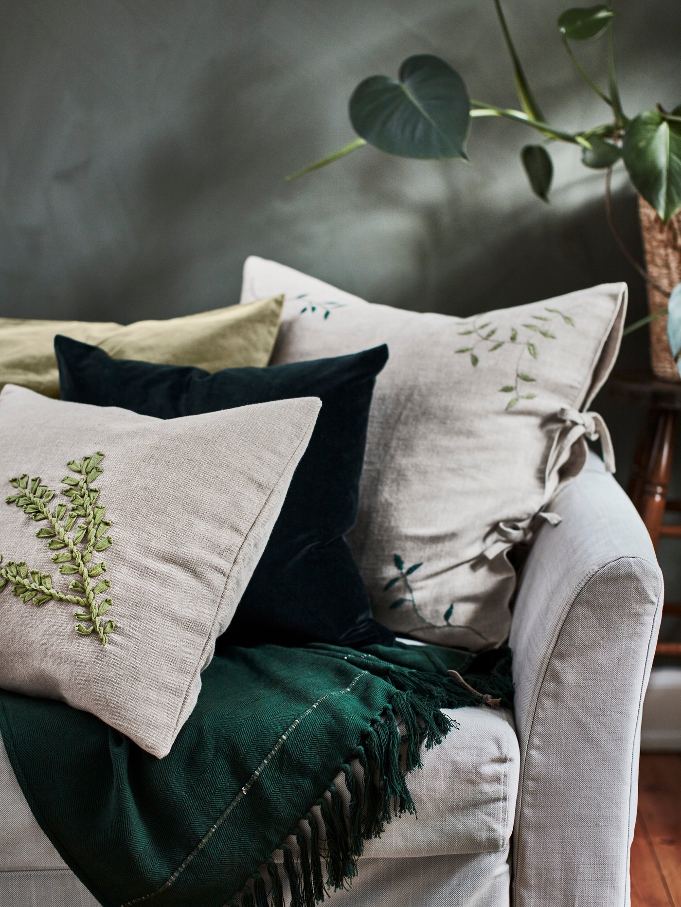 One green and three beige cushions decorated with unique leaf embroideries on a light grey sofa with a dark green throw.