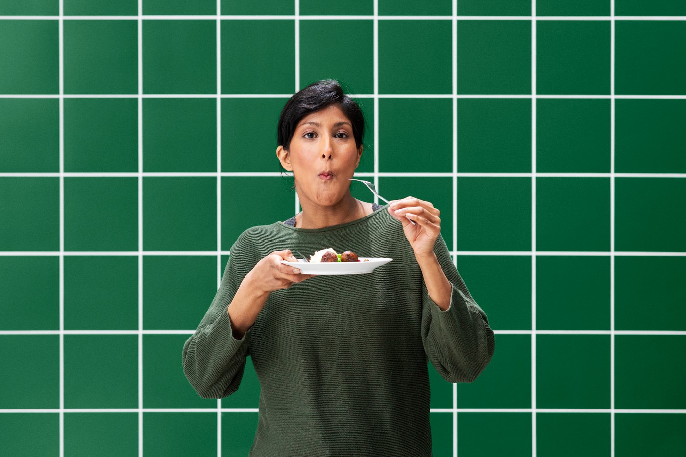 A woman is holding a plate of plant balls and has a fork up to her mouth tasting one.