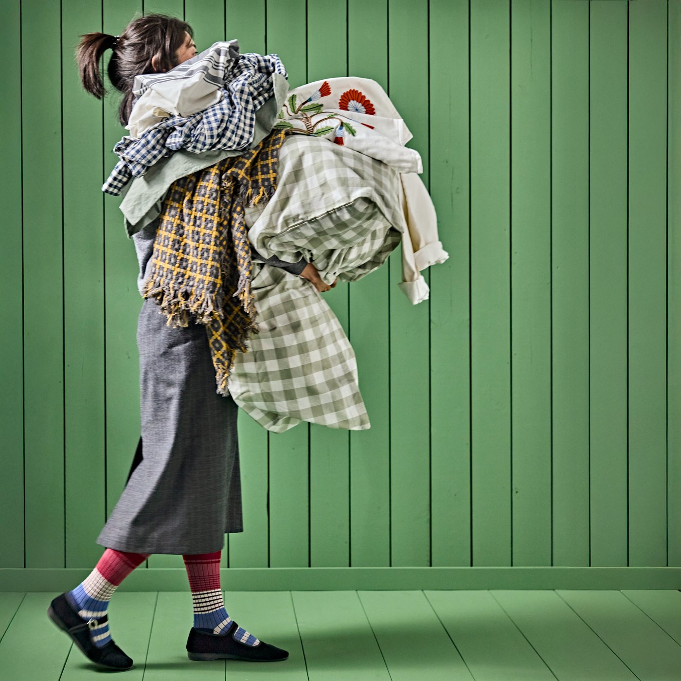 A persona carrying a large bundle of clothes and textile products against a green wall and floor of colour green.