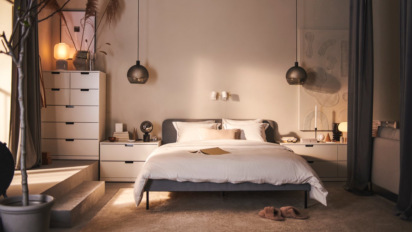 A SLATTUM upholstered bed and various white NORDLI chests of drawers stand against a wall in a bedroom.