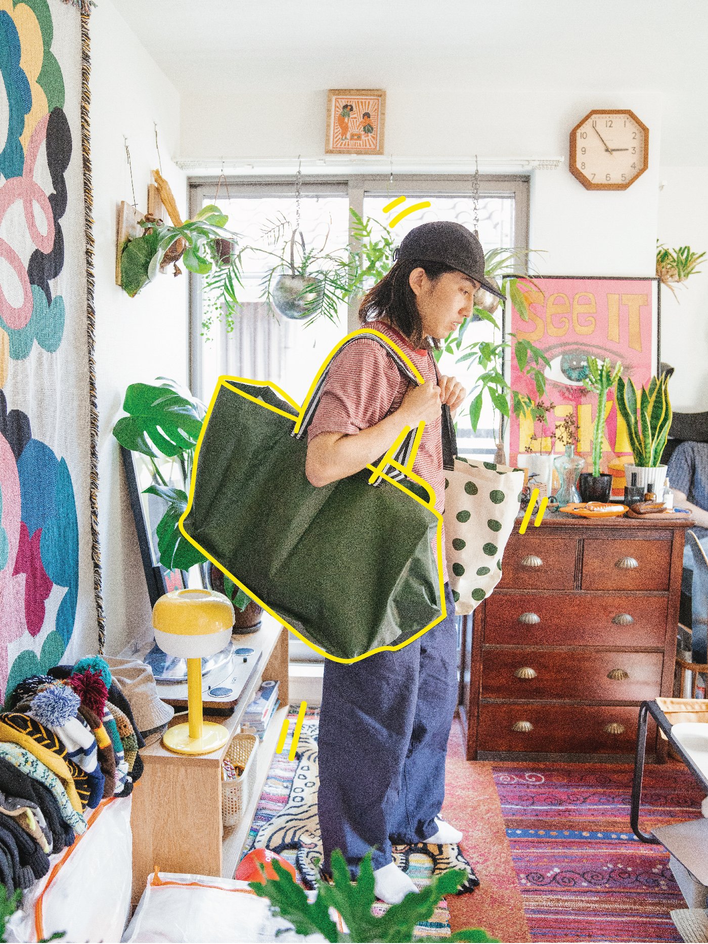 Eine Person steht in einem hellen Raum und hält eine grosse grüne Tragetasche und eine weisse Tasche mit Polka-Punkten in den Händen, umgeben von Pflanzen und bunten Dekorationen.