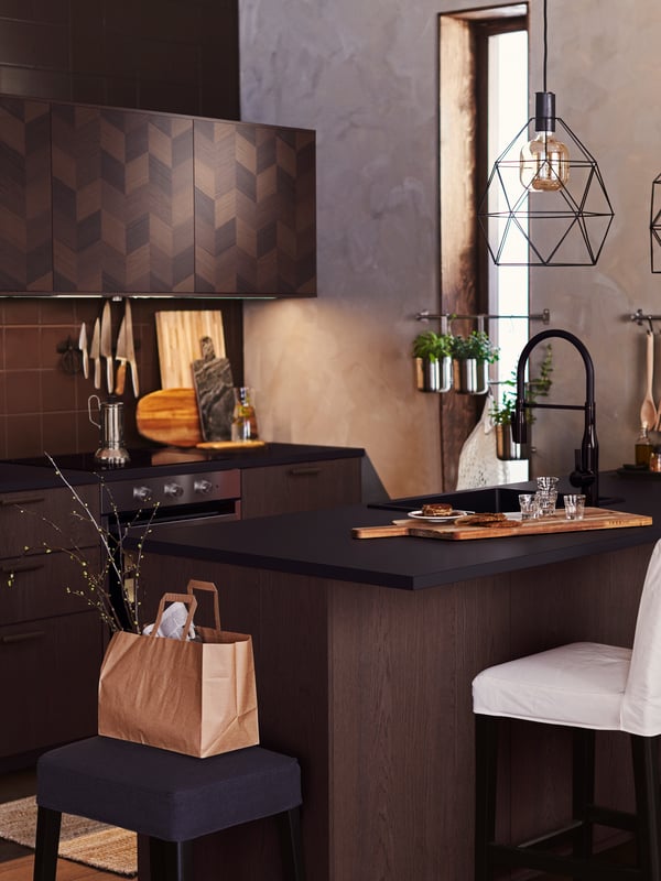 A dark wood kitchen with an island that has a BRUNSTA pendant lamp overhead and glasses on the counter.