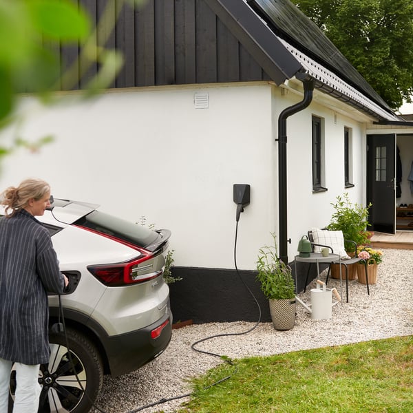 Une voiture électrique est en train de se recharger à l'extérieur d'une maison moderne. Une borne de recharge est fixée au mur. On aperçoit également de la verdure, comme des plantes et des arbres.