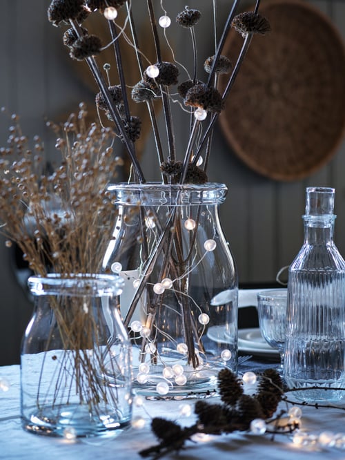 Gros plan d’un vase BEGÄRLIG en verre clair contenant des fleurs séchées, des branches et une guirlande lumineuse à DEL SNÖYRA sur une table festive.