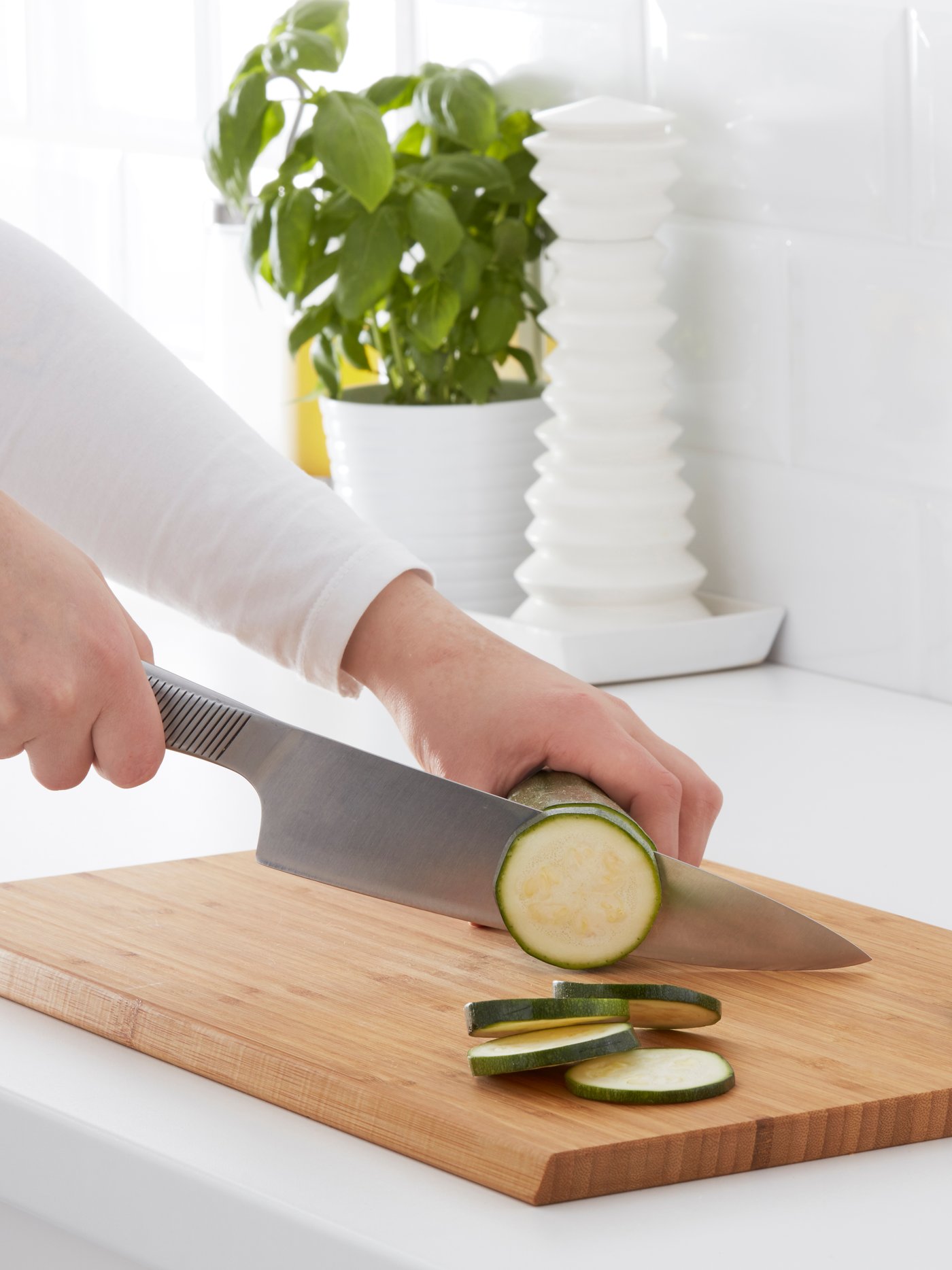 The hands of a person using APTITLIG bamboo chopping board and a large kitchen knife to slice a zucchini.