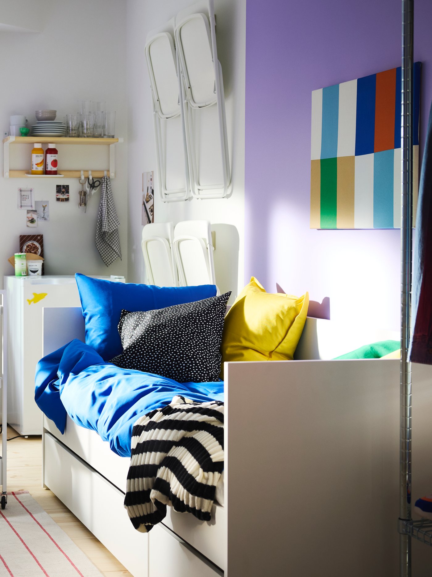 Sovrum med stora fönster med lila målade bågar, en vit dagbädd, en rullvagn och en grön/vit ÖNNESTAD fåtölj.