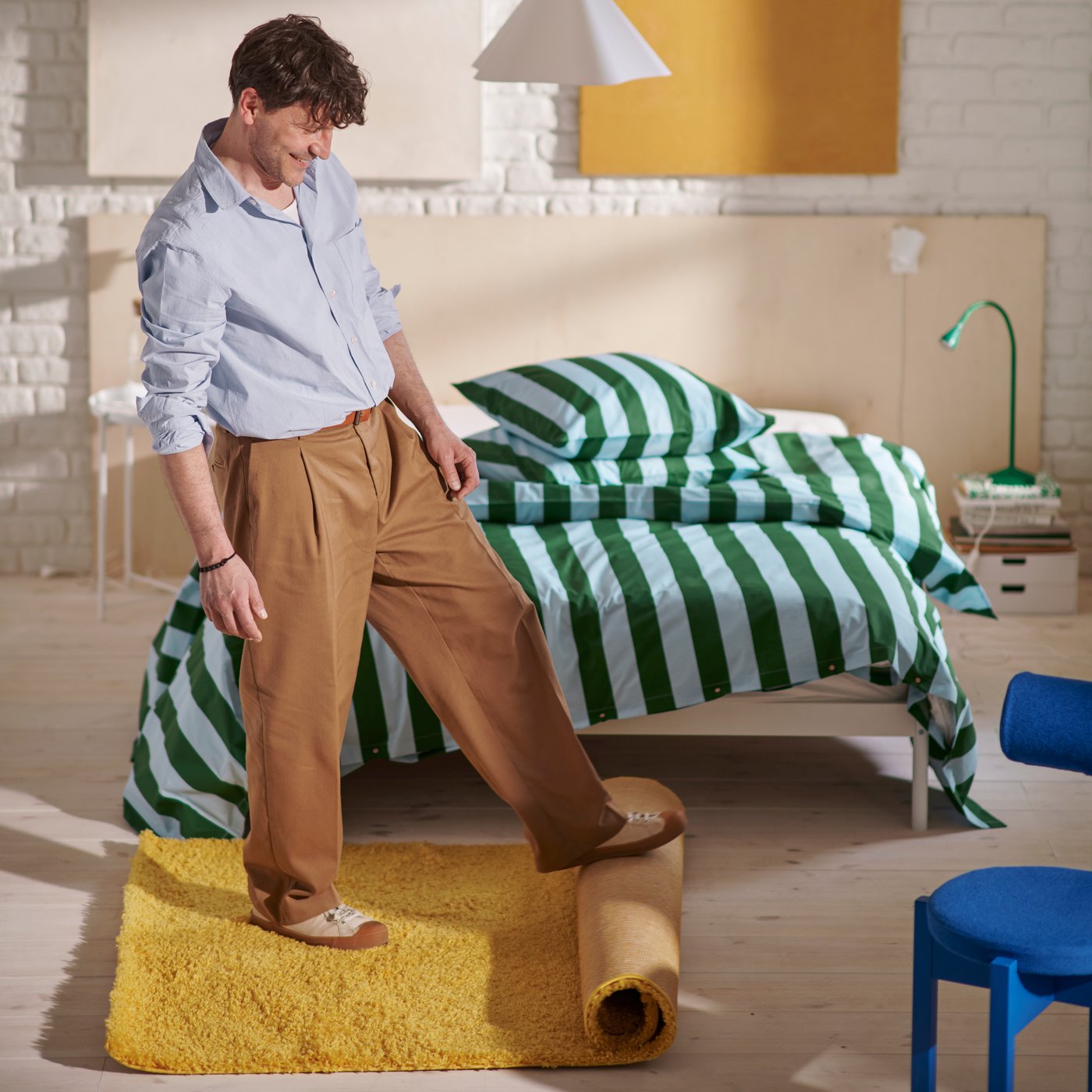 A man rolls out a yellow ÄRENDE rug in a bedroom with a wooden bed and blue and green striped SLÅNHÖSTMAL bedding.