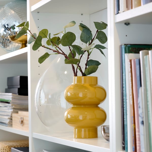 A yellow-beige PÄRLHÄGG vase holding leafy branches, placed in a BILLY bookcase framed by books and decoration.