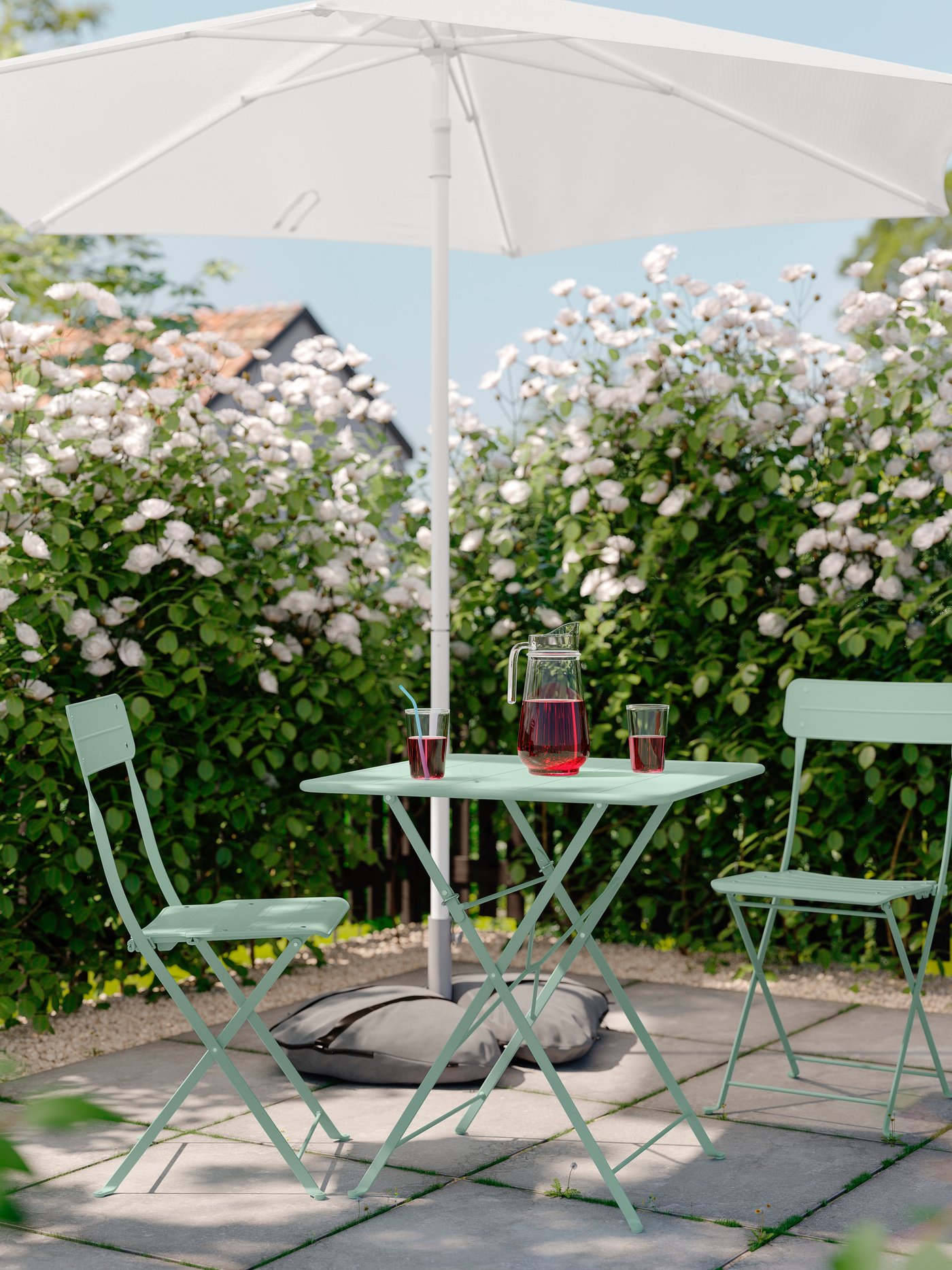 Ett mintgrönt caféset med två stolar och runt bord under parasoll i en blommande trädgård.