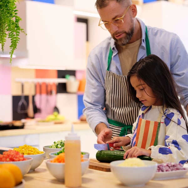 A grown-up is helping a child cut vegetables in a kitchen. There are many white IKEA 365+ bowls with veggies on the worktop.