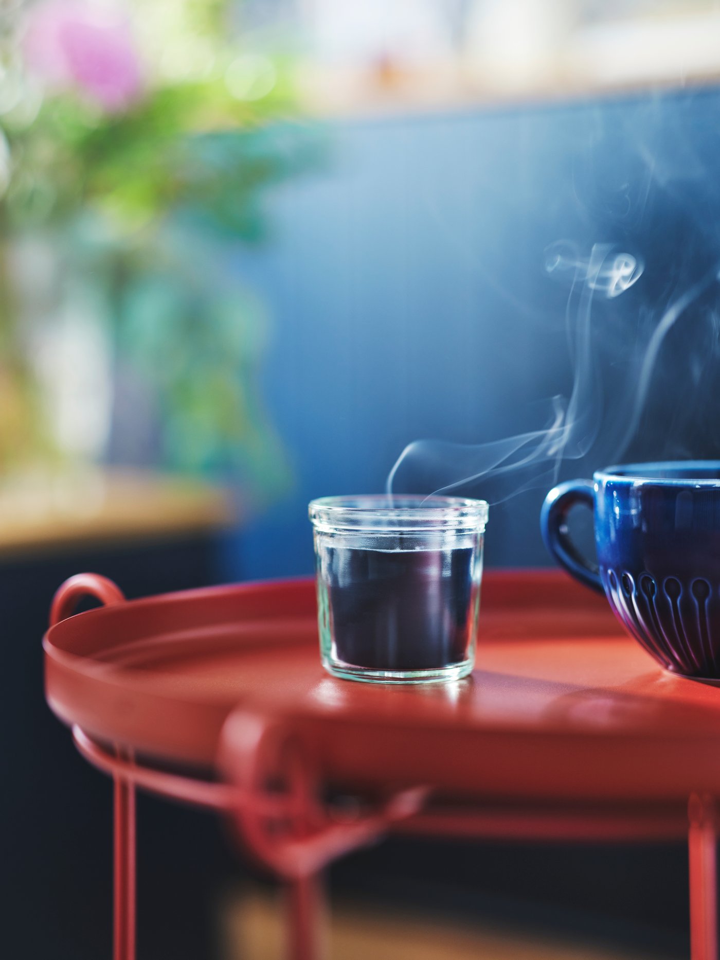 Eine FRUKTSKOG Duftkerze im Glas steht neben einem blauen STRIMMIG Becher aus Steinzeug auf einem roten LIVELYCKE Tabletttisch.