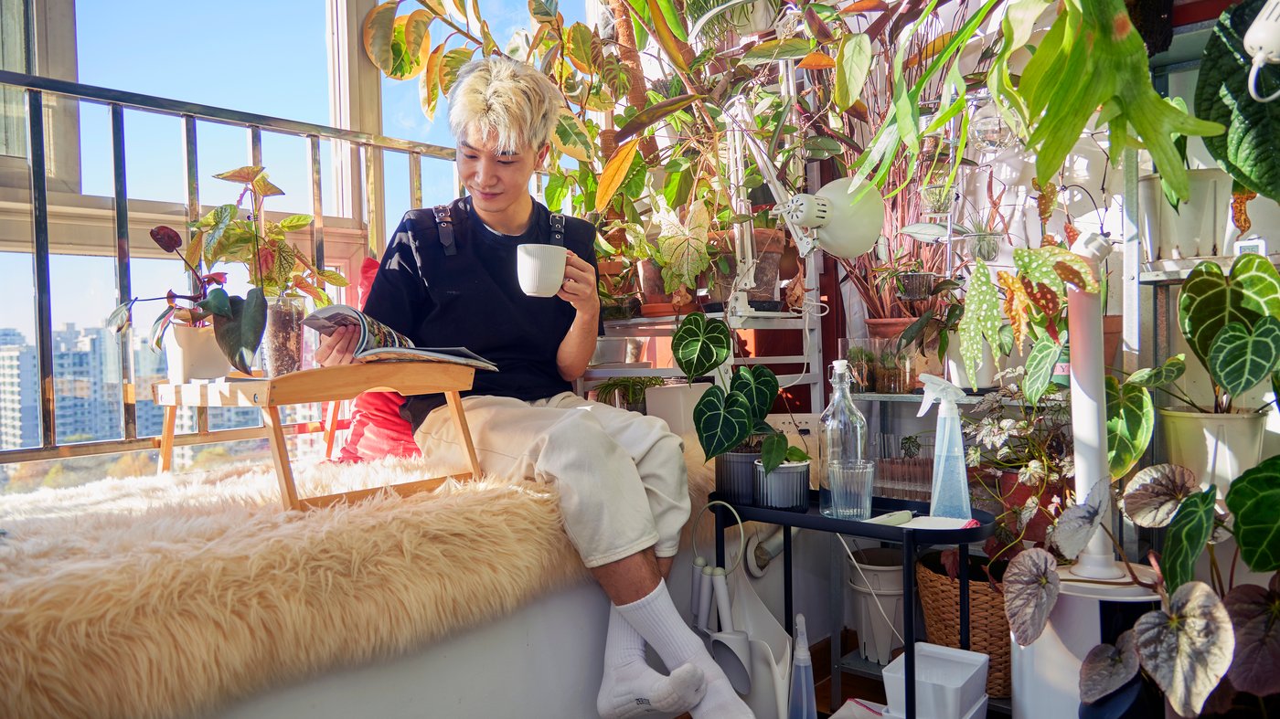 Man sitting next to a window, reading a book and drinking coffee, surrounded by house plants.