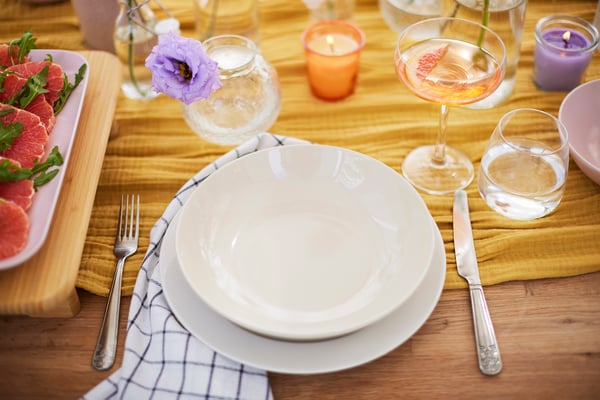 Una tavola imbandita composta da un piatto basso e uno profondo, una coppa per lo champagne, un bicchiere per l'acqua e posate eleganti in rilievo. Sul tavolo, con un runner al centro, ci sono vasi con fiori e candele profumate in un bicchiere di vetro.