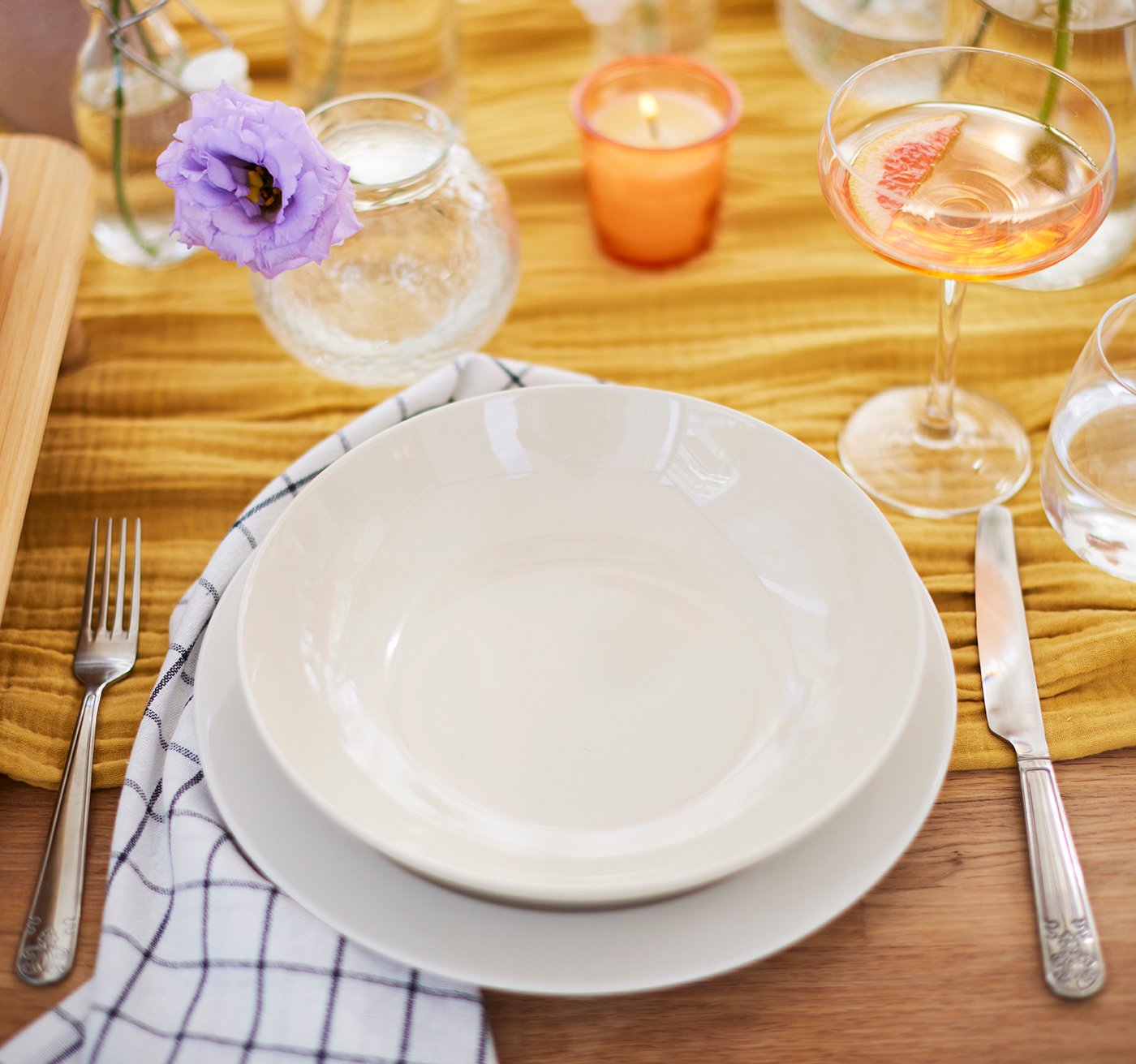 Ein Tischgedeck aus flachem und tiefem Teller, einer Sektschale, Wasserglas und elegantem Besteck mit Relief. Auf dem Tisch mit Tischläufer in der Mitte stehen Vasen mit Blumen und Duftkerzen im Glas