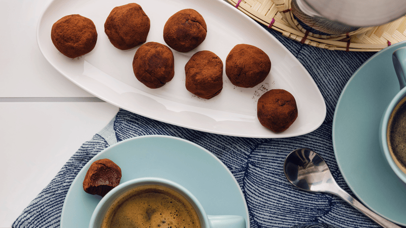 Wit bord in de vorm van een blad waarop zeven chocoladetruffels liggen naast twee turkooizen kopjes met schoteltje waar koffie in zit.