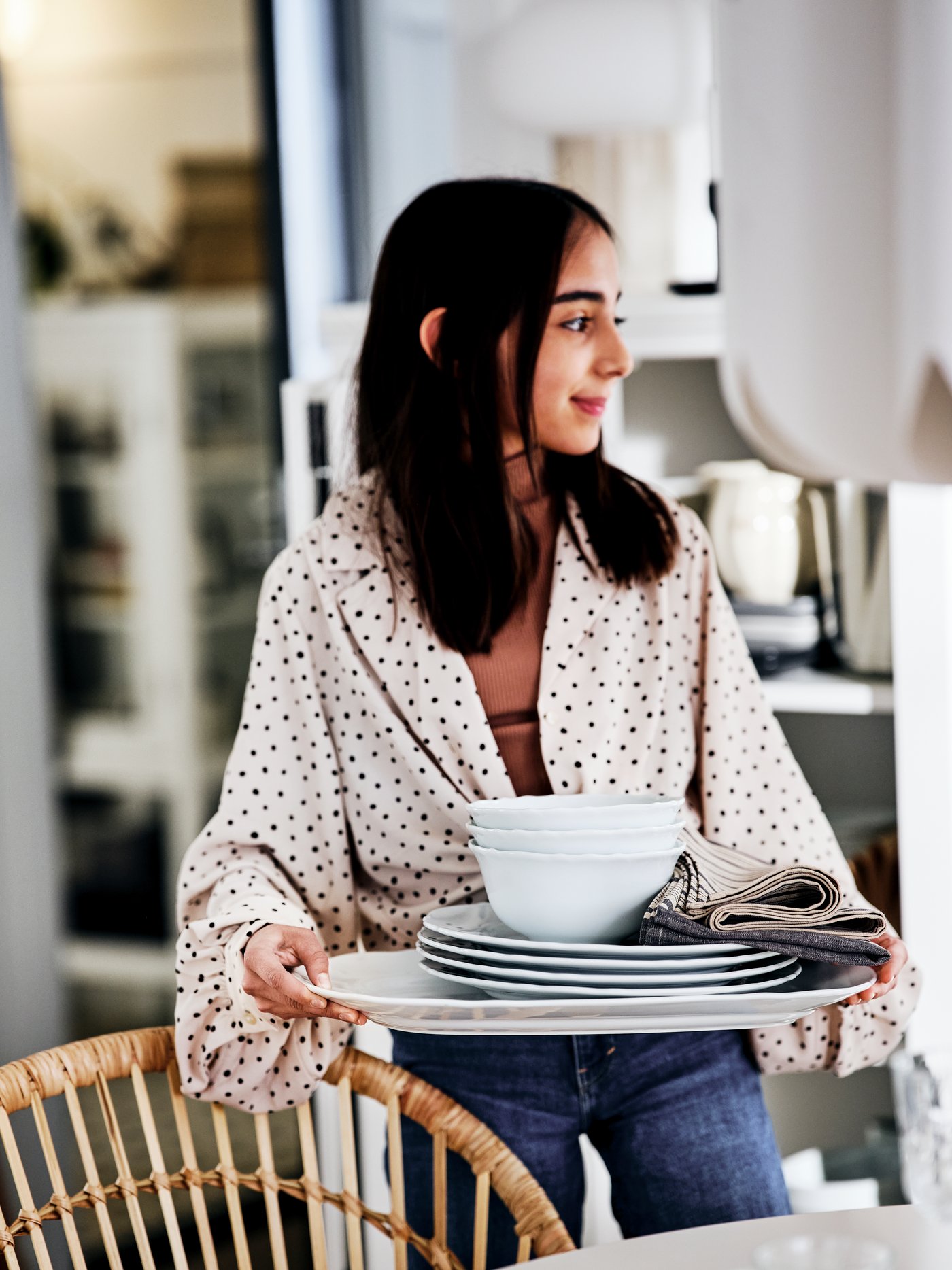 A young woman holding a tray with plates and bowls on it beside a table and a NILSOVE rattan armchair.
