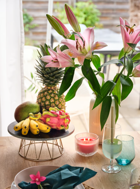 Tischarrangement mit tropischen Früchten, rosa Lilien in Vase, Kerze und Glas auf hell gedecktem Tisch am Fenster.